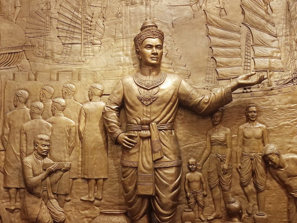 Bas-relief doré montrant un roi couronné, bras tendu, entouré de courtisans, de villageois et de jonques à voiles.