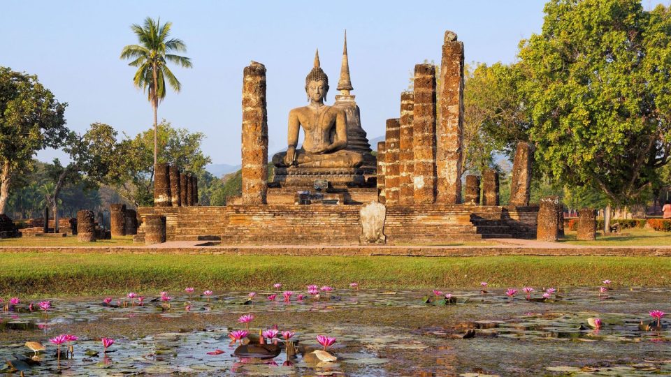 Un Bouddha assis dans la posture de la prise de la terre à témoin, encadré par les colonnes d'un ancien wihan et adossé à un chedi en bouton de lotus, forme caractéristique de l'art de Sukhothai. — © Asia King Travel Bouddha assis en briques et stuc entre des colonnes en ruine, devant un chedi, avec un étang de nénuphars roses au premier plan.