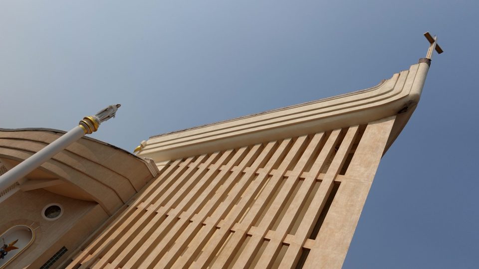 La cathédrale Saint-Michel-Archange de Tha Rae : la façade en claustra s'élève en pointe, comme une proue de bateau, jusqu'à la croix sommitale. — © Eric Monteil Façade beige en contre-plongée, à claustra de lames croisées, surmontée d'une croix, à côté d'une statue sur colonne blanche.