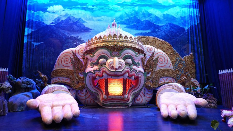 Décor de scène du centre d'apprentissage du khon de Bang Pa-in : une figure monumentale de yak couronné, dont la bouche éclairée sert d'entrée aux danseurs. — © Eric Monteil Scène de théâtre ornée d'une tête géante de démon couronné, mains ouvertes, bouche éclairée, devant un décor de montagnes bleues.
