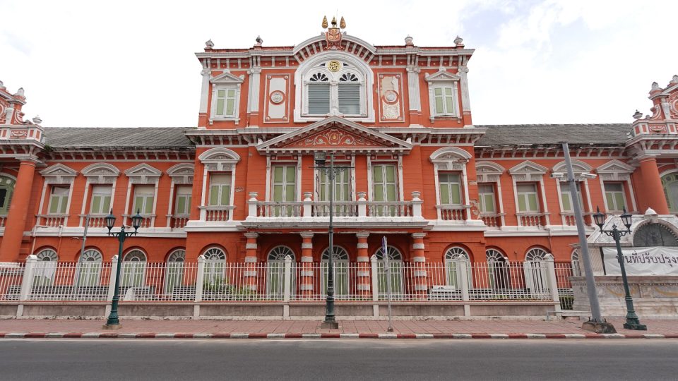 Le bâtiment du Département royal de topographie, à Phra Nakhon : une façade symétrique d'inspiration européenne, avec avant-corps central à fronton, balcon à balustres et fenêtres cintrées soulignées de stuc blanc. — © Eric Monteil Façade d'un bâtiment de style européen en briques rouge orangé, avec stucs blancs, volets verts et grille, au bord d'une rue.