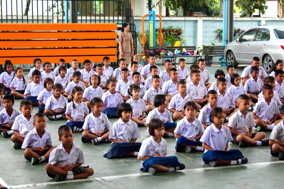 Rassemblement d'élèves d'une école primaire thaïlandaise : chemisette blanche brodée de l'écusson de l'établissement, short ou jupe bleu marine, assis en rangs sous un préau. — © ThailandeFR Des dizaines d'écoliers en uniforme blanc et bleu assis en tailleur sur un sol couvert, une enseignante debout derrière eux.