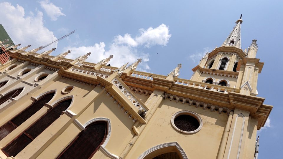 Façade néogothique crème d'une église vue en contre-plongée, avec clocher à flèche, pinacles et fenêtres en arc brisé sous un ciel bleu.