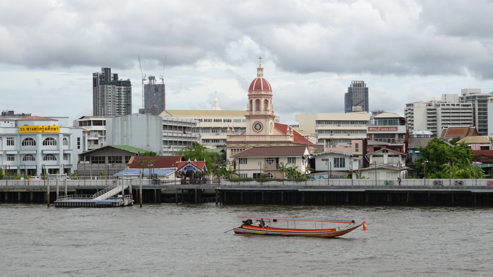 L'église Santa Cruz vue depuis le Chao Phraya, avec sa tour-lanterne surmontée d'un dôme rouge et d'une croix, dominant les quais de Kudi Chin ; au premier plan, un bateau à longue queue (ruea hang yao). — © Eric Monteil Église au dôme rouge et clocher crème sur une berge de fleuve, bateau à longue queue au premier plan, immeubles derrière.