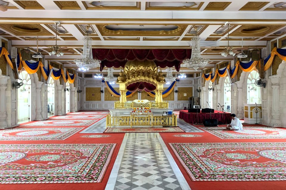 Grande salle de prière sikhe avec tapis rouges à motifs floraux, lustres en cristal et dais doré central abritant le livre saint.