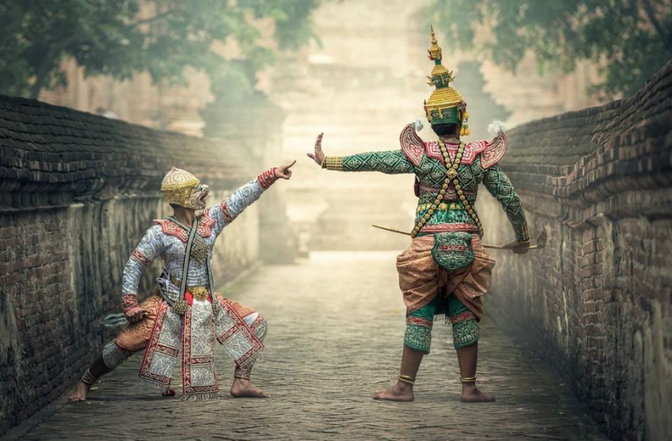 Deux personnages du khon, théâtre dansé masqué : dans cette tradition, chaque posture des mains, des bras et des jambes porte un sens codifié. — © Mysakonnakhon.com Deux danseurs en costumes de khon, l'un masqué en singe blanc, l'autre en vert coiffé d'une couronne pointue, s'affrontent dans une allée pavée entre deux murs de briques.