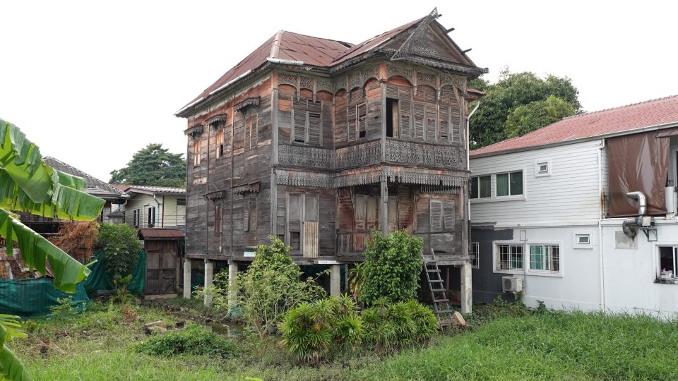 Une maison en bois à Kudi Chin, élevée sur piliers maçonnés : remarquer les lambrequins et balustrades ajourés, les persiennes et le pignon décoré, aujourd'hui très dégradés. — © Eric Monteil Maison ancienne en bois à deux étages sur pilotis, aux frises ajourées, au toit de tôle rouillée, entourée de végétation.