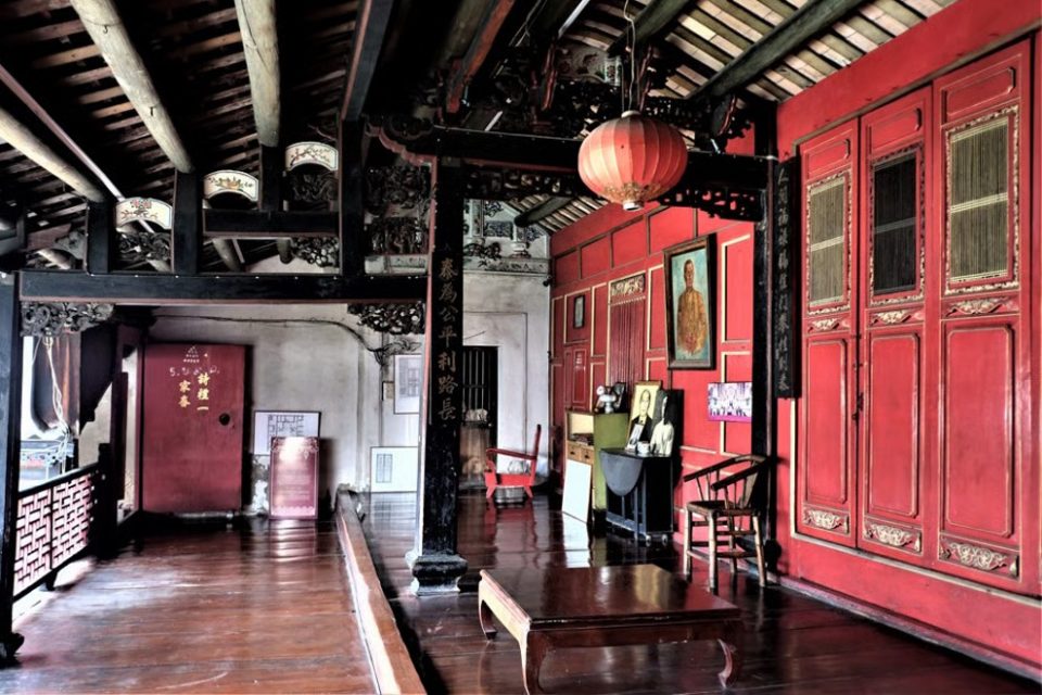 Intérieur d'une demeure de style chinois : poutres sculptées, cloisons de bois laquées de rouge et inscriptions verticales en caractères chinois sur les piliers. — © South China Morning Post Intérieur d'une maison chinoise ancienne à charpente sombre sculptée, murs et portes laqués rouges, lanterne rouge et portrait encadré.