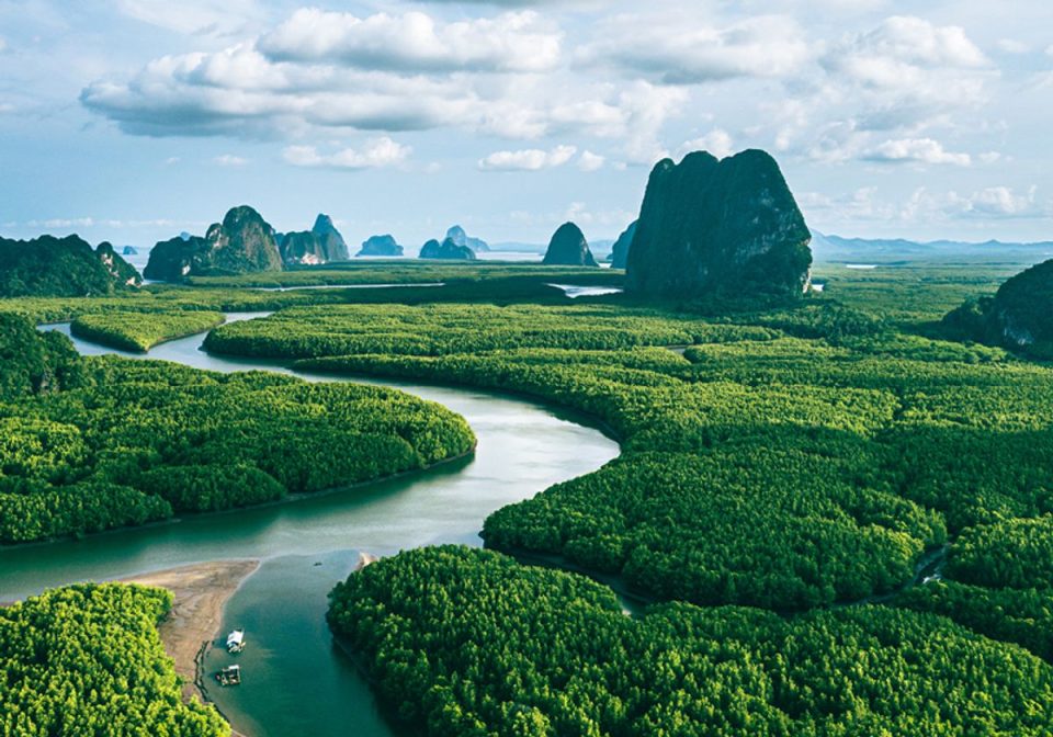 Une rivière à marée serpente dans une mangrove continue ; au loin, des pitons karstiques émergent de la plaine et de la mer. — © The Scientist Vue aérienne d'une rivière sinueuse traversant une mangrove dense, avec des pitons calcaires à l'horizon et deux petits bateaux.