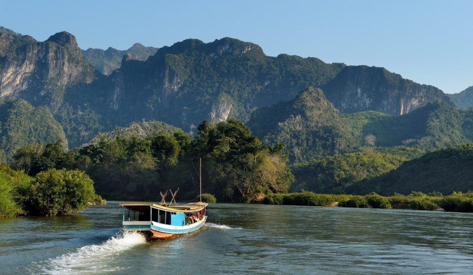 Un bateau en bois bleu et blanc à toit long remonte un large fleuve brun-vert, au pied de montagnes karstiques boisées.