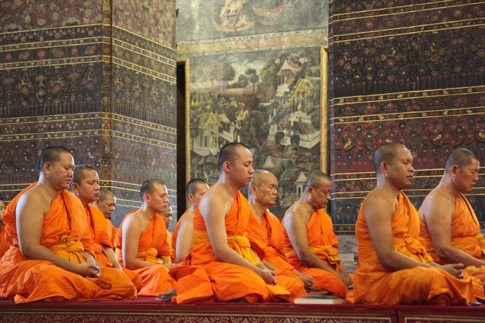 Moines bouddhistes en méditation dans une salle de wat ; à l'arrière-plan, piliers ornés de motifs floraux et peinture murale figurant des pavillons à toits étagés. — © Newmandala.org Moines en robes orange assis en rangée, yeux mi-clos, devant des piliers peints de motifs floraux et une peinture murale.