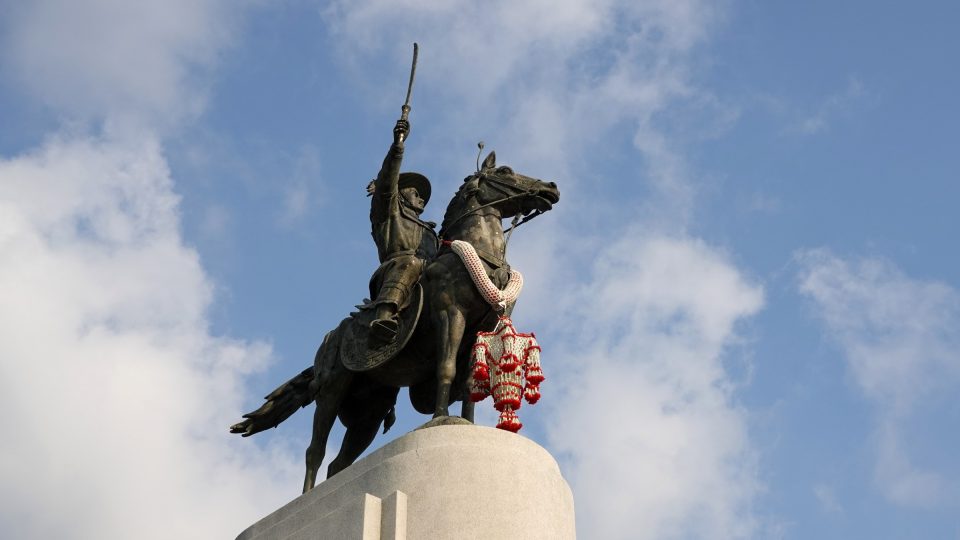 Monument du roi Taksin le Grand à Thonburi : le souverain, épée levée, chevauche sur un haut socle ; les guirlandes de fleurs témoignent des offrandes qui lui sont faites. — © Eric Monteil Statue équestre en bronze du roi Taksin brandissant une épée, une guirlande de fleurs au cou du cheval, sur socle de pierre claire.