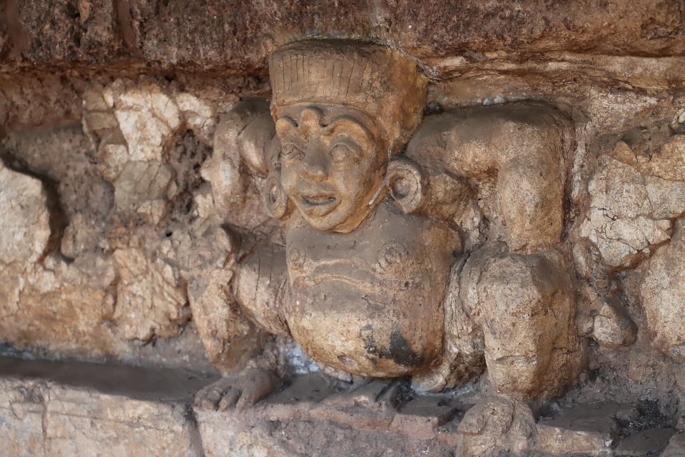 Figure de nain porteur (gana) au visage rond et à la coiffe cylindrique, accroupi sous une corniche dont il semble soutenir le poids, motif fréquent dans le décor des soubassements de monuments bouddhiques anciens. — © Salaween.blog Figure sculptée en stuc ou pierre, accroupie, bras levés, coiffe haute et grands ornements d'oreilles, soutenant une corniche.