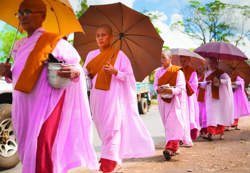 Des nonnes bouddhistes en robe rose et étole safran partent en procession, bol à aumônes en main ; ces vêtements ne correspondent pas à la tenue blanche des mae chi de Thaïlande. — © The Buddhist Review File de nonnes bouddhistes au crâne rasé, en robes roses et étoles orangées, marchant sous des parapluies avec des bols.
