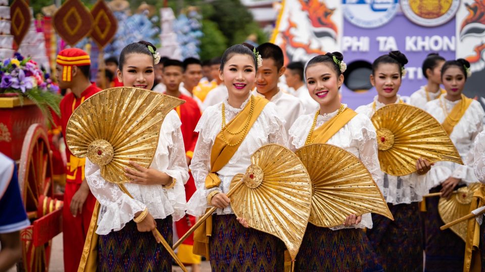 Danseuses en costume traditionnel, éventails dorés en main, dans le cortège du festival Phi Ta Khon ; une banderole porte le nom de la fête à l'arrière-plan. — © Amazing Thailand Jeunes femmes en chemisier de dentelle blanche et jupe tissée, tenant de grands éventails dorés lors d'un défilé.