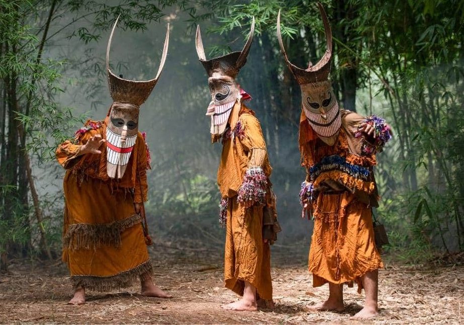 Trois Phi Ta Khon en costume : masques aux yeux ronds et longues dents, surmontés d'une coiffe en vannerie à cornes recourbées, et tenues frangées de pompons colorés. — © Odasie Trois personnes masquées, coiffes à longues cornes et costumes orange à franges, posent pieds nus dans un bosquet de bambous.