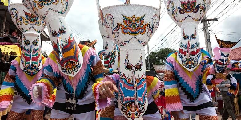 Défilé des Phi Ta Khon à Dan Sai : les masques, faits d'un panier à riz gluant en guise de coiffe et d'une base façonnée dans une gaine de palmier, se prolongent d'un long nez peint ; les costumes sont cousus de bandes de tissu colorées. — © Asia King Travel Participants costumés portant de grands masques peints aux longs nez et coiffes hautes, en vêtements multicolores à franges, dans une rue.