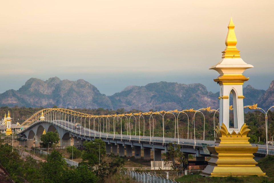 Un pont routier franchit la vallée du Mékong ; ses lampadaires dorés en forme d'ondulation et les colonnes ornementales blanc et or annoncent un passage frontalier, devant les reliefs calcaires de la rive laotienne. — © Jinnyz.com Long pont routier aux arches et lampadaires dorés, avec une colonne blanche et dorée au premier plan et des montagnes karstiques au fond.