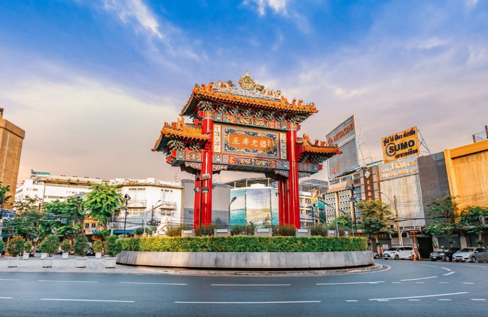 La porte de style chinois du rond-point d'Odeon marque l'entrée du Chinatown de Bangkok : piliers rouges, toits superposés aux tuiles vernissées et panneau central portant une inscription en caractères chinois. — © Hotel Ambert Bangkok Grande porte chinoise rouge à toits dorés au centre d'un rond-point planté de haies, entourée d'immeubles et d'enseignes.