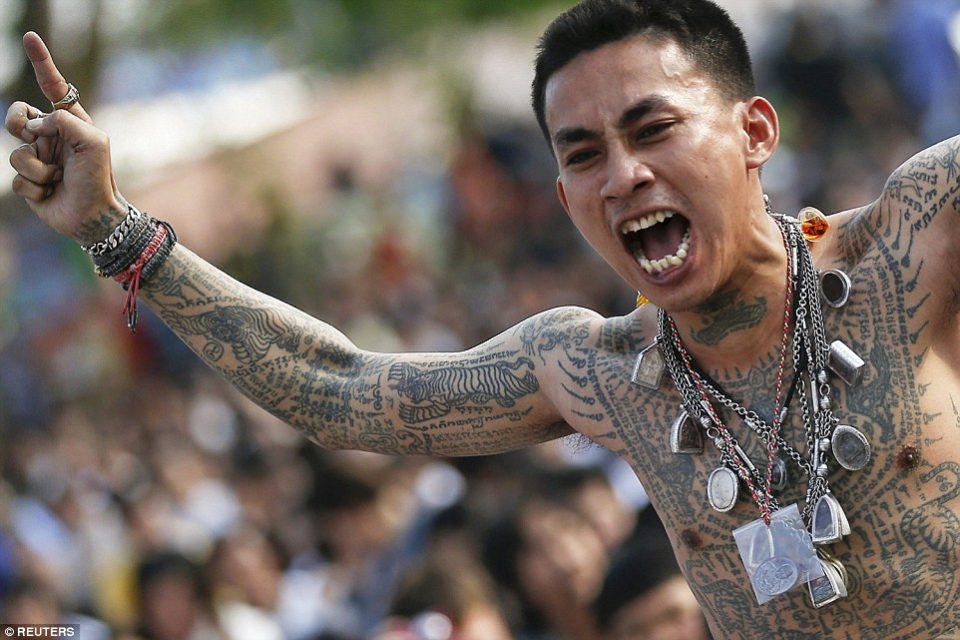 Un homme tatoué de sak yant, dont un tigre sur le bras et des grilles de caractères sur le torse, porte de nombreuses amulettes en pendentif. — © Reuters Homme torse nu couvert de tatouages sacrés, amulettes au cou, criant le bras levé devant une foule.