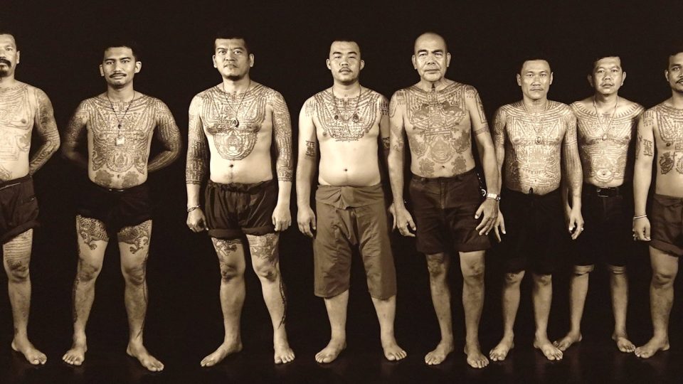 Hommes portant des sak yant sur le torse, les bras et les jambes : grilles de caractères, figures protectrices et motifs géométriques couvrent presque tout le corps. — © Eric Monteil Huit hommes torse nu alignés pieds nus sur fond noir, le torse, les bras et les jambes couverts de tatouages.