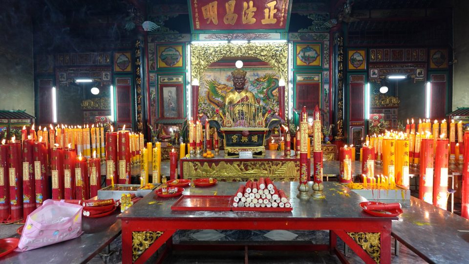 Le sanctuaire de Chow Sue Kong à Talat Noi : devant l'autel de la divinité, les fidèles allument de grands cierges rouges et jaunes, tandis que des paquets de bougies attendent sur la table d'offrandes. — © Eric Monteil Intérieur d'un sanctuaire chinois : statue dorée d'une divinité sur un autel, entourée de grands cierges rouges et jaunes allumés.