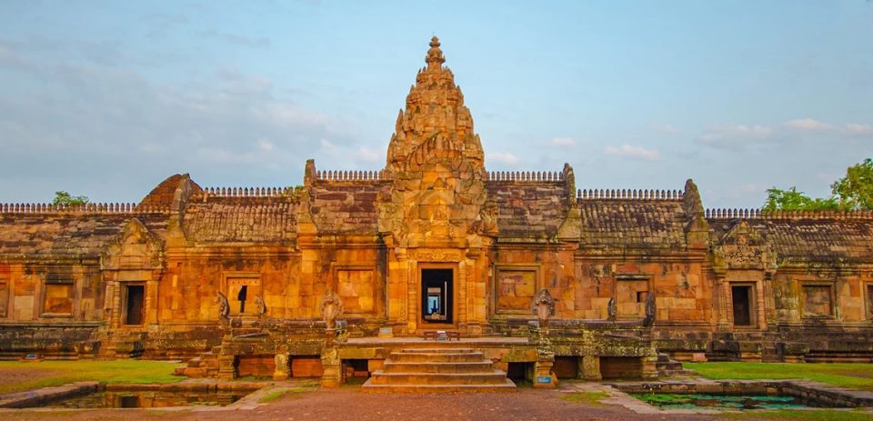 Un sanctuaire khmer en grès : la tour centrale (prasat) se dresse au-dessus des galeries qui l'entourent, avec un escalier axial menant à l'entrée. — © Grantourismotravels.com Sanctuaire khmer en grès rose avec une tour centrale ouvragée, des galeries à toits de pierre et un escalier d'accès entre deux bassins.