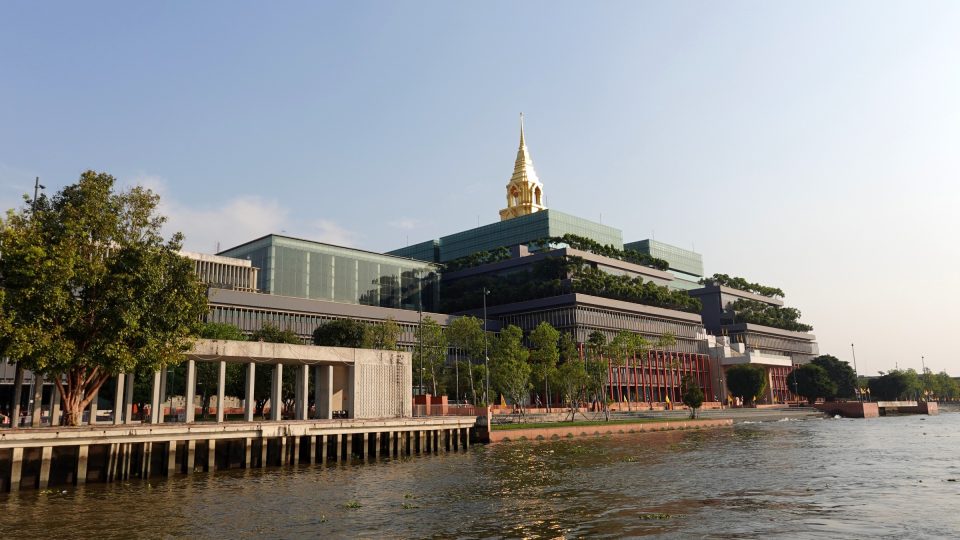 Le Sappaya-Sapasathan, siège du Parlement thaïlandais au bord de la Chao Phraya, dans le district de Dusit : terrasses plantées étagées et flèche dorée inspirée du chedi. — © Eric Monteil Grand bâtiment moderne à terrasses végétalisées surmonté d'une flèche dorée, vu depuis un fleuve, avec quai et arbres au premier plan.