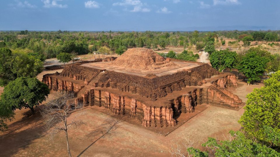 Grand monument bouddhique en latérite de la période de Dvaravati : un soubassement carré à redans et escaliers axiaux porte les restes arasés d'un chedi. — © Salaween.blog Vue aérienne d'un grand monument en latérite à plan carré et terrasses, surmonté d'un tertre de briques, entouré d'arbres.