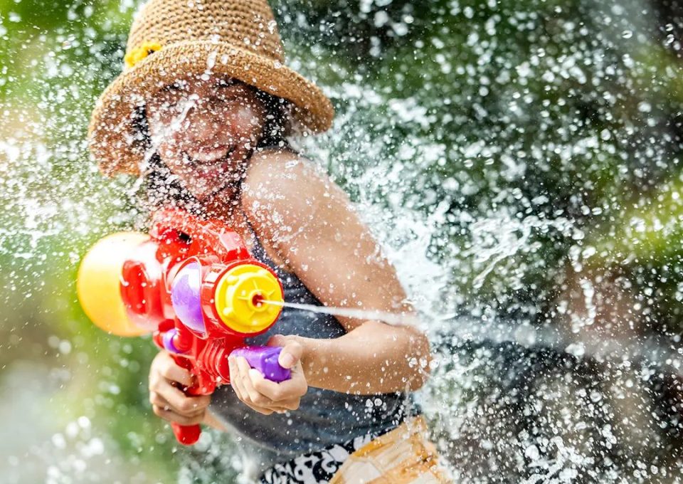 Bataille d'eau au pistolet en plastique, une scène typique des réjouissances de Songkran, le Nouvel An thaï d'avril. — © Amazing Thailand Une femme coiffée d'un chapeau de paille tire avec un pistolet à eau en plastique coloré, entourée d'éclaboussures.