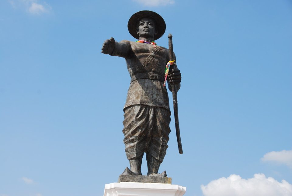 Statue du roi Anouvong de Vientiane, bras tendu et sabre en main, ornée de guirlandes et de rubans laissés en offrande ; ce souverain lao se révolta contre le Siam en 1826-1828. — © Flickr Statue en bronze d'un homme coiffé d'un chapeau, bras droit tendu, tenant un sabre, sur fond de ciel bleu.
