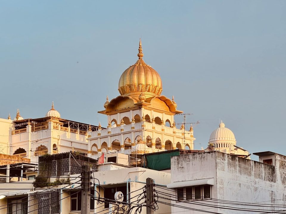Le dôme doré du grand temple sikh de Pahurat domine les immeubles et les enchevêtrements de câbles du quartier. — © 8riwroy Dôme doré en forme de bulbe surmontant un édifice blanc à arcades, au-dessus de toits et de fils électriques.