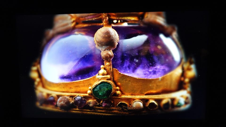 Pièce d'orfèvrerie présentée parmi les trésors d'or issus de la crypte du wat Ratchaburana : une pierre violette en cabochon, enchâssée dans l'or et rehaussée d'une émeraude et d'une rangée de petites gemmes. — © Eric Monteil Gros plan d'un objet en or serti d'une grande pierre violette polie, d'une émeraude et de petites gemmes, sur fond noir.