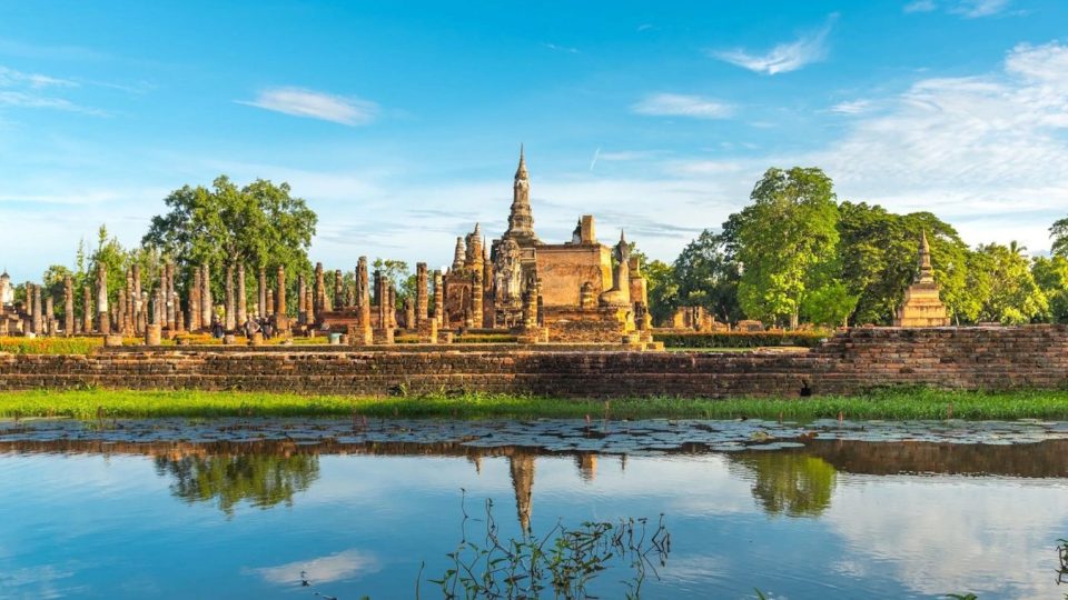 Le wat Mahathat de Sukhothai : derrière le bassin, les colonnes du wihan mènent au chedi central couronné d'un bouton de lotus, forme propre à l'architecture de Sukhothai. — © Eric Monteil Ruines de temple en briques avec chedi en bouton de lotus, rangées de colonnes et bassin reflétant le ciel bleu.