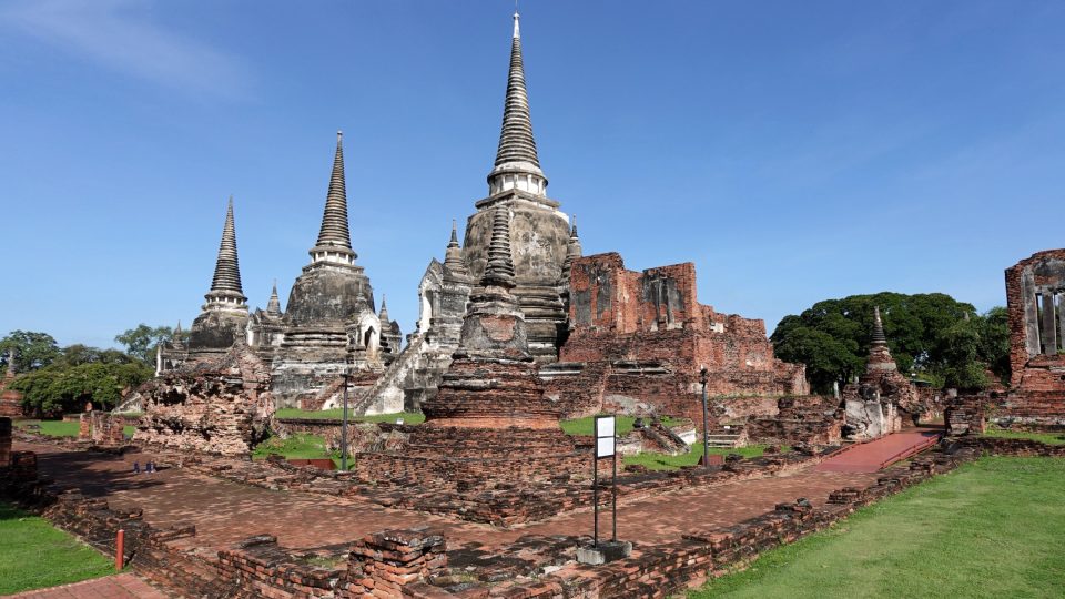 Trois grands chedi en forme de cloche, gris et élancés, dominant des ruines de briques rouges sous un ciel bleu.