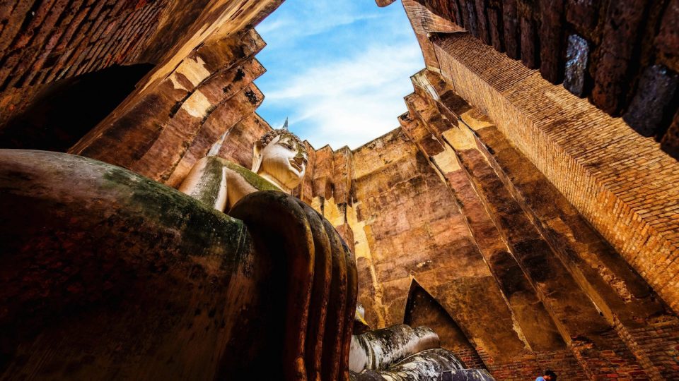 Bouddha monumental dans un mondop de brique à ciel ouvert : les doigts effilés de la main posée sur le genou, au premier plan, relèvent du style de Sukhothai. — © Amazing Thailand Grand Bouddha assis en stuc blanc vu en contre-plongée entre de hauts murs de brique ouverts sur le ciel, main posée au premier plan.
